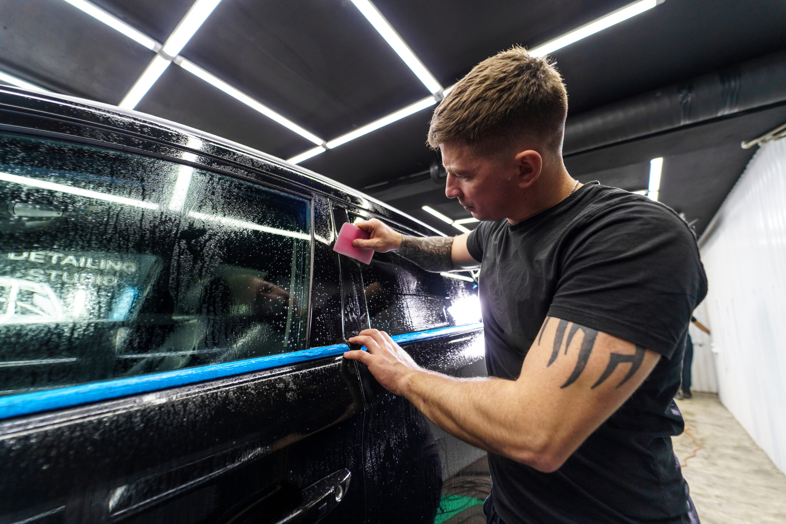 A tattooed man applies protective film to a black car at an auto detailing studio